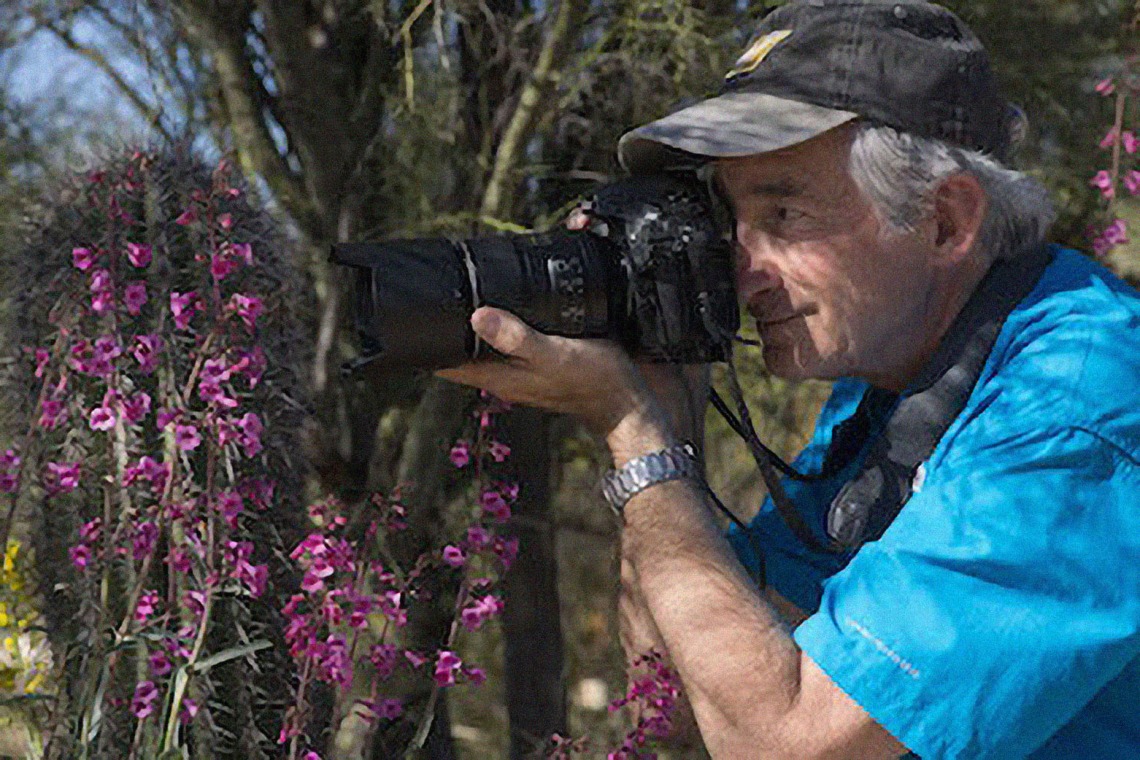 Stephen Buchman in a desert landscape, pointing a camera toward a subject to the left of the frame.
