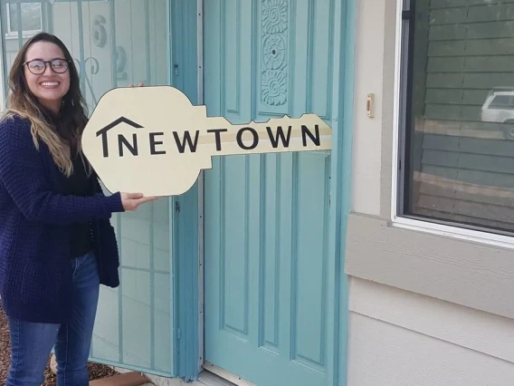 A smiling woman stands outside of a residential dwelling holding a key-shaped sign imprinted with the words NEWTOWN.