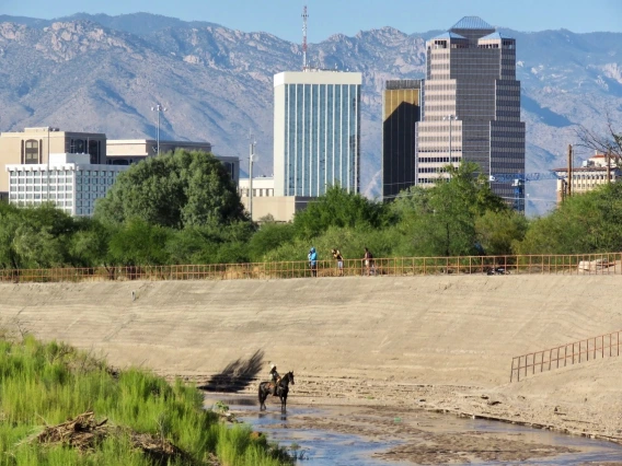 Someone riding a horse in the middle of the santa cruz river with downtown tucson buildings rising above in the background