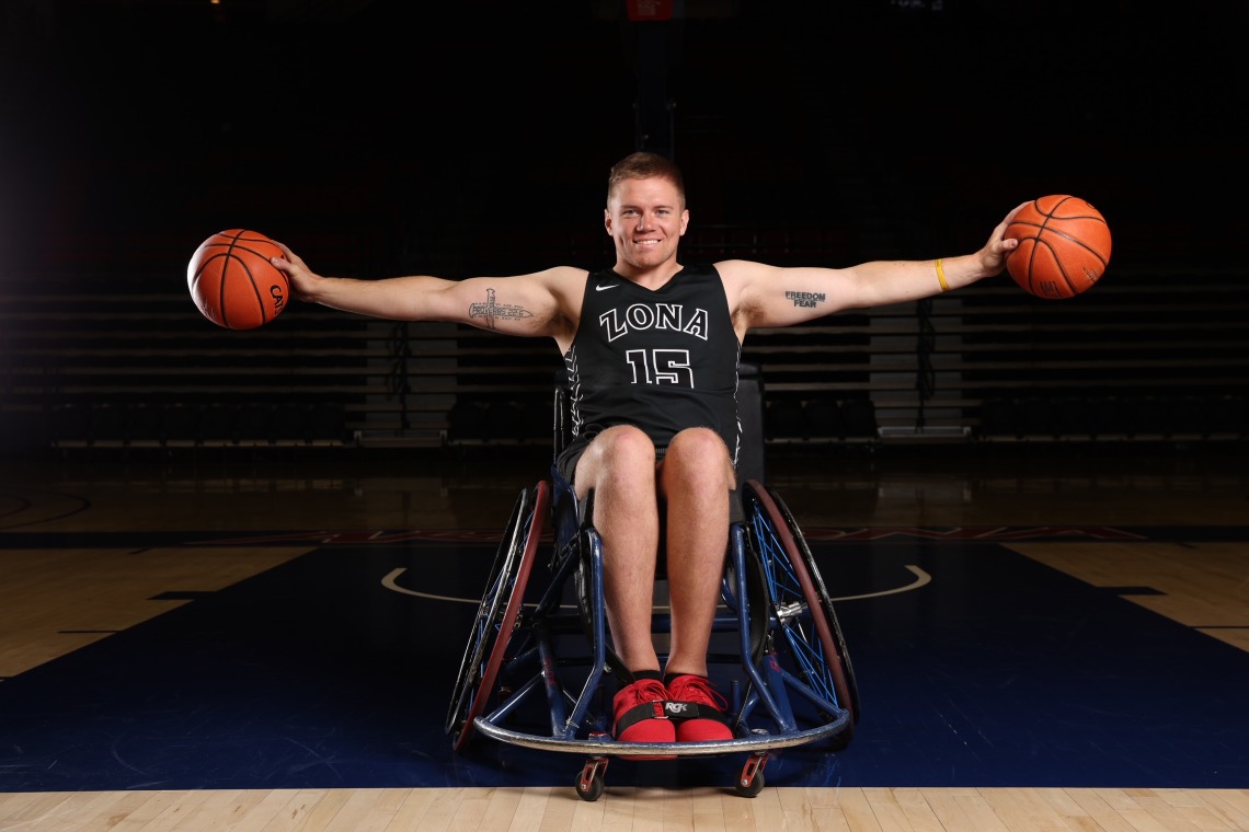 Hunter Pinke, an man using a wheelchair, holds two basketballs in both hands outstretched.  