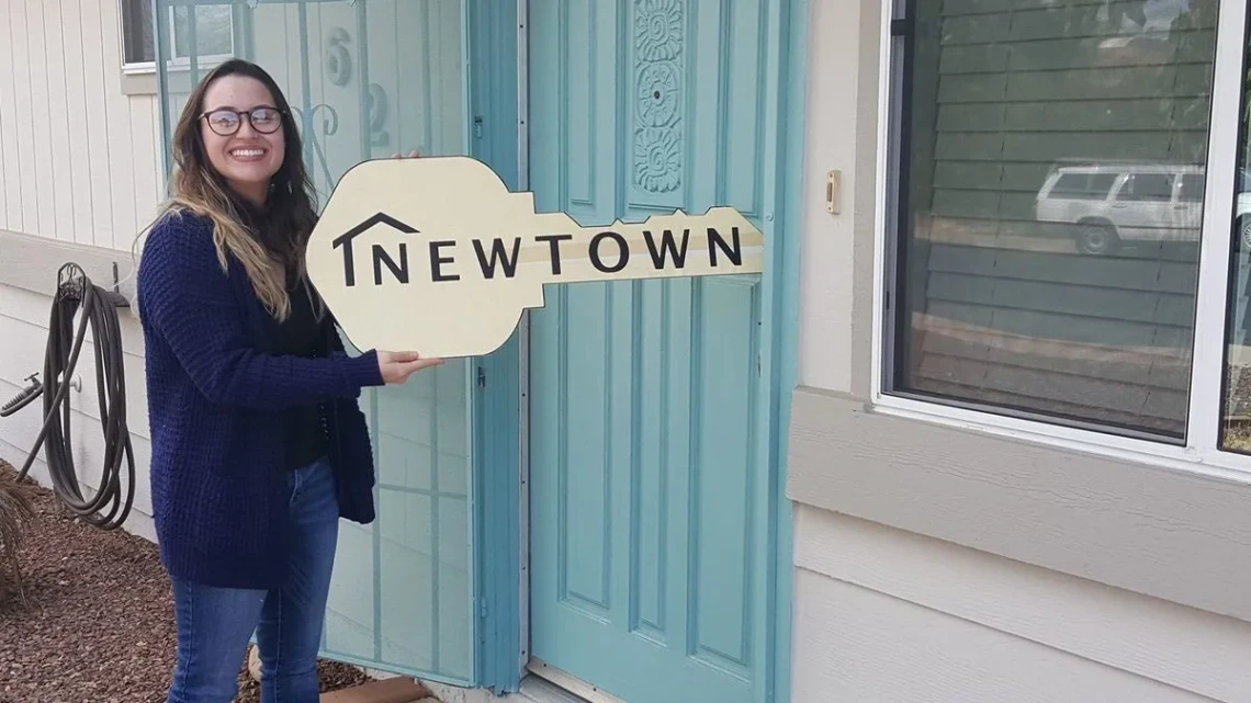 A smiling woman stands outside of a residential dwelling holding a key-shaped sign imprinted with the words NEWTOWN.