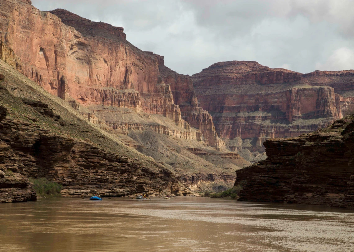 View of the Colorado river from the bottom of the Grand Canyon: two small rafts can be seen on the water in the distance and high cliffs rise in the background